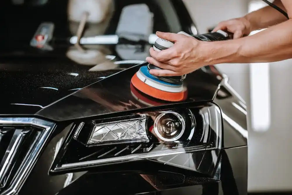 Detailer using a dual-action polisher on a black car's hood and headlight during paint correction and polishing